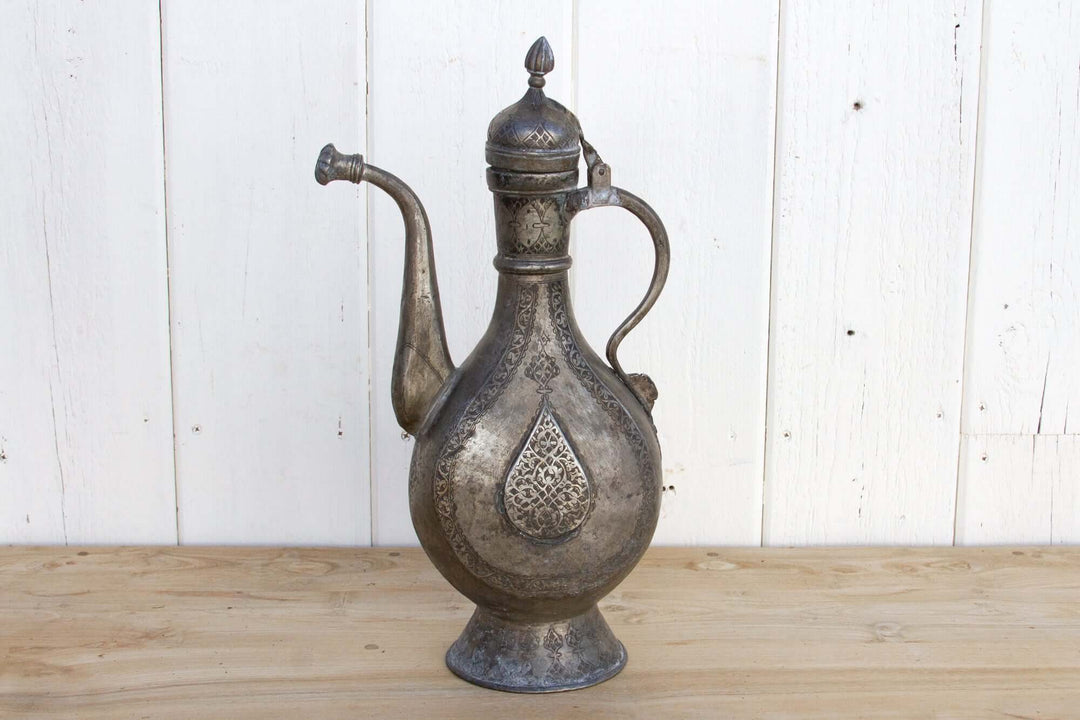 Decorative metal pitcher with intricate designs on a wooden surface and white wooden panel background