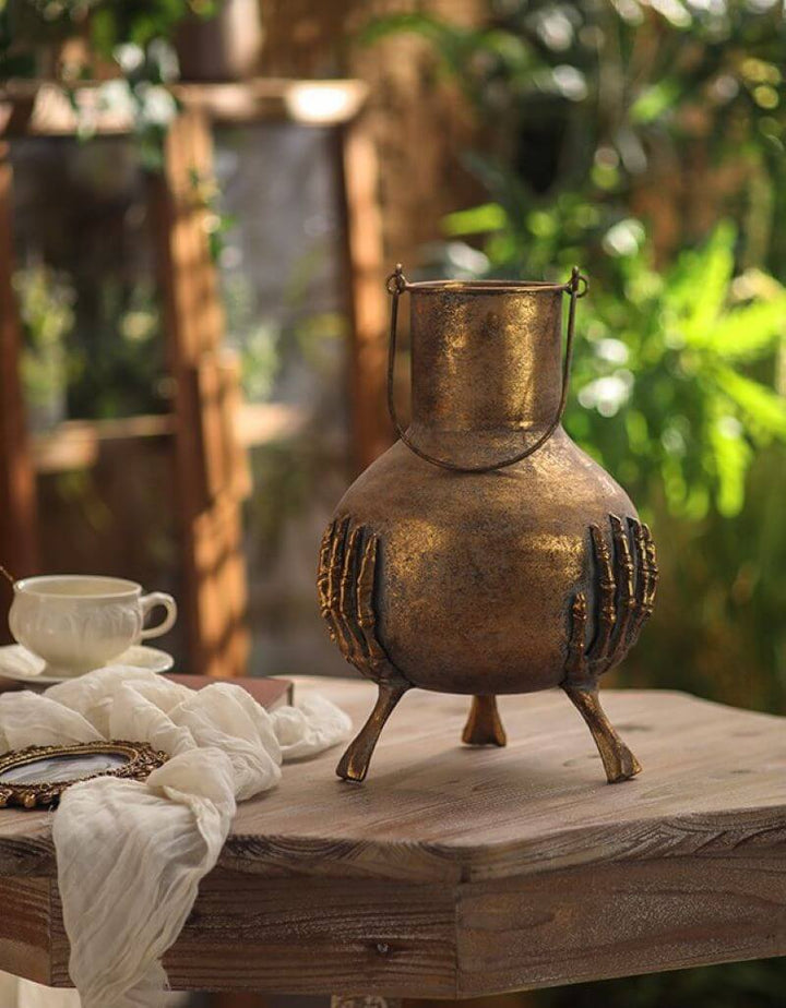 Gothic Skeleton Hands Iron Vase with antique gold finish on a wooden table, surrounded by greenery and a teacup.