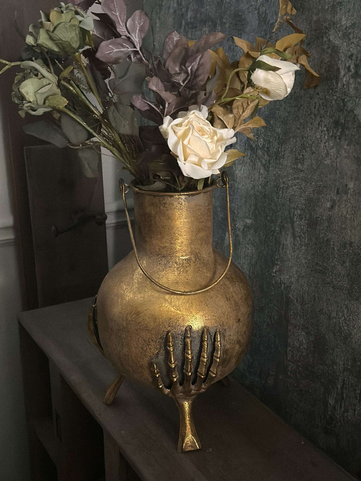 Gothic Skeleton Hands Iron Vase with antique gold finish, held by three skeleton hands, filled with flowers, on wooden shelf.