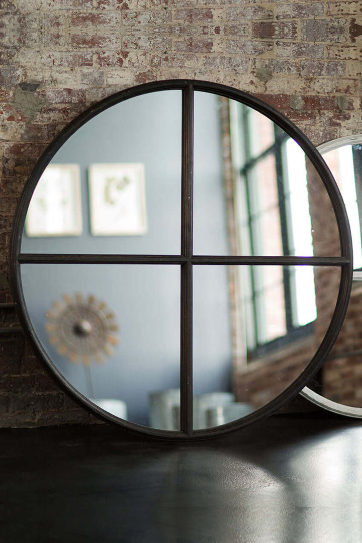 Large round wood wall mirror in black with crossbar design against a textured brick wall.