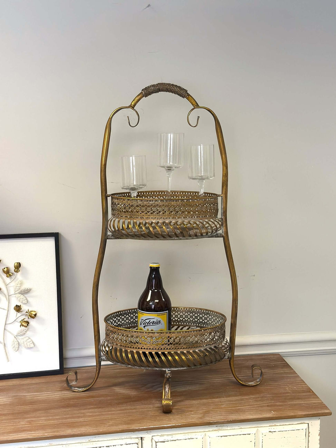 Vintage brass 2-tier metal bar cart with ornate detailing, holding wine glasses and a bottle, on a wooden surface.