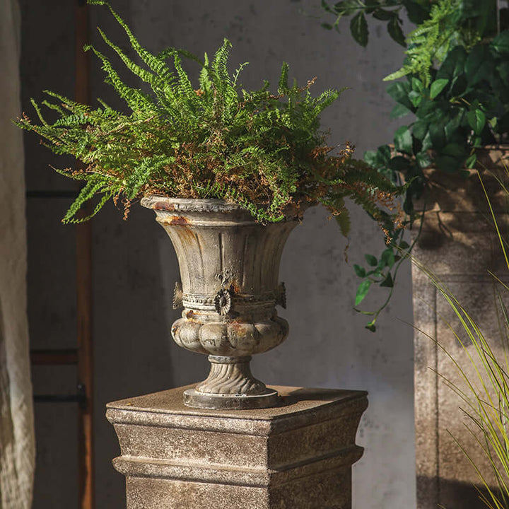 Decorative urn with a fern on a stone pedestal against a gray wall.