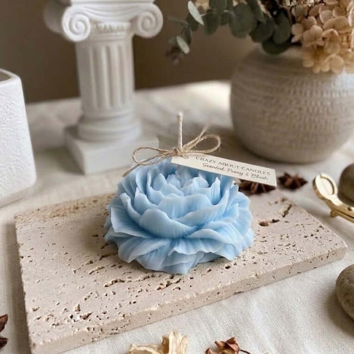 A light blue flower-shaped candle wrapped in twine with a tag reading 'Crazy Candle Co. Sacred Flow & Air,' styled on a stone slab with decorative props including a column, vase, and dried flowers.