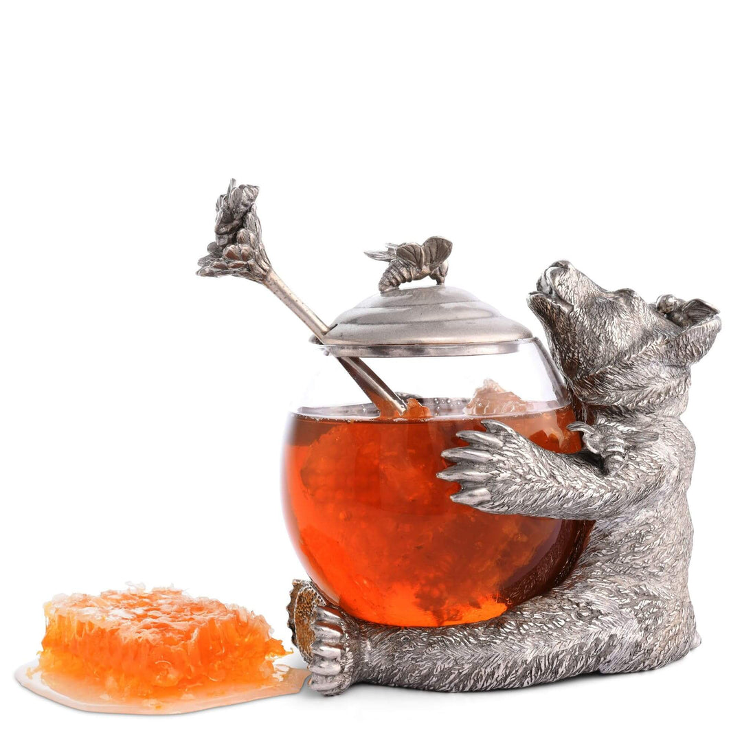 Decorative jar with silver bear holding a glass container filled with honey on a white background.