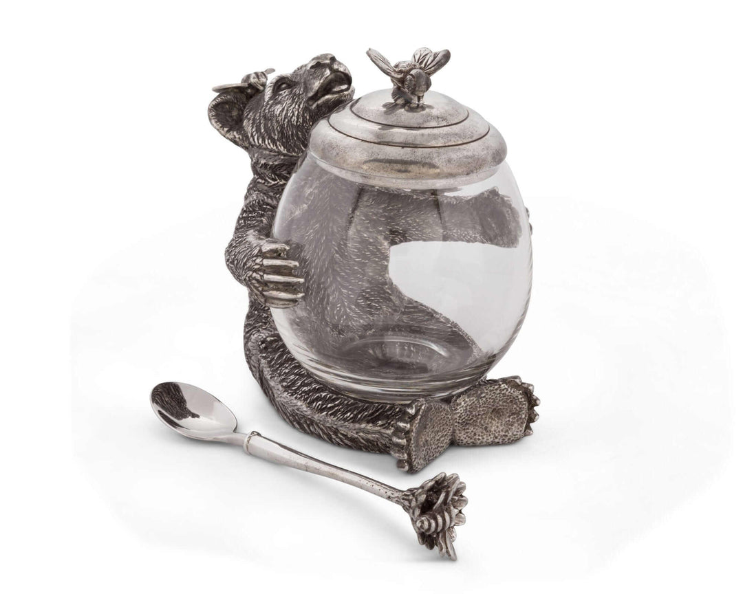 Decorative glass jar with metal bear and bee design, accompanied by a spoon on a white background.