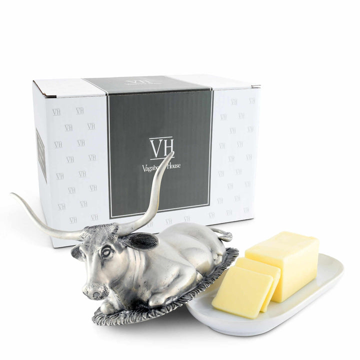 Metallic cow-shaped butter dish with butter and a branded box on a white background