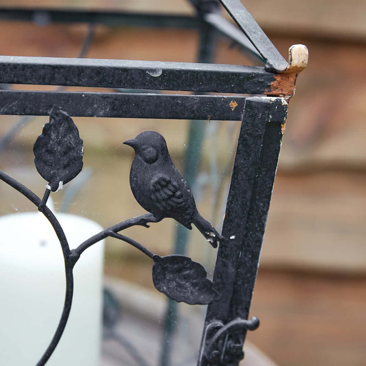 Close-up of a rustic lantern design featuring a bird and leaf motif on the metal frame.
