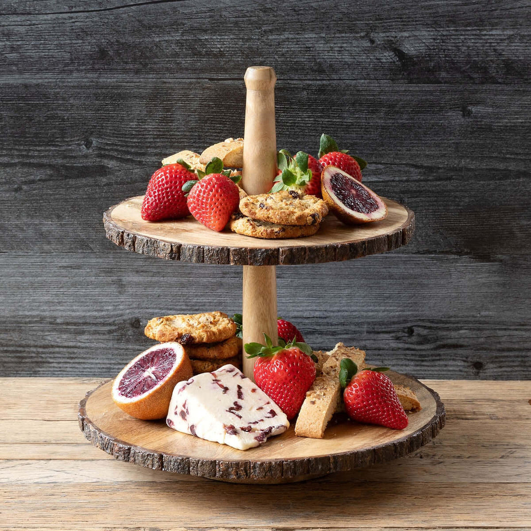 Two-tiered wooden cake stand with fruits, cheese, and cookies on a wooden surface.
