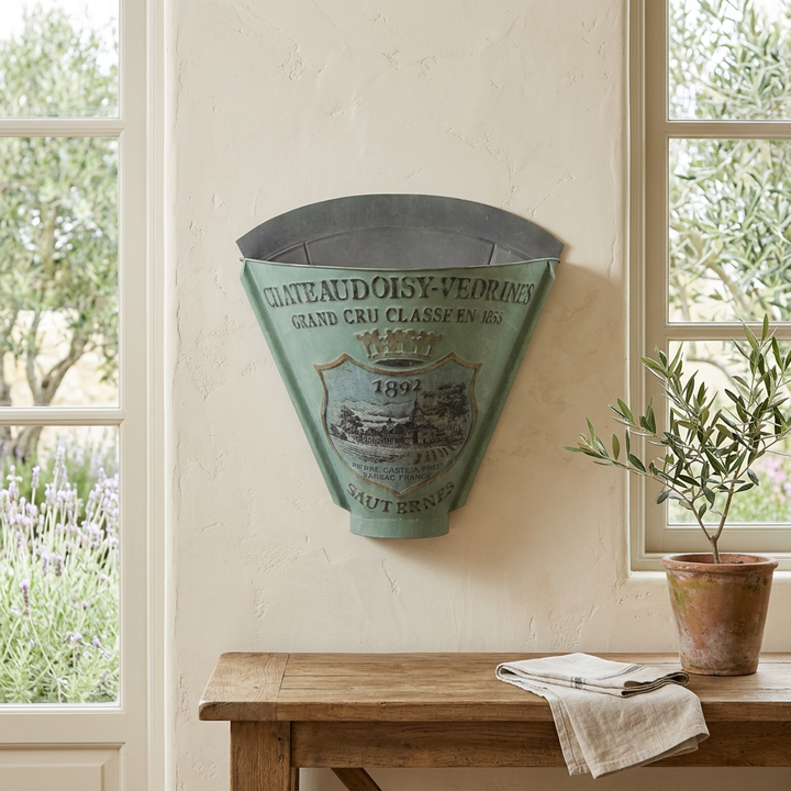 Sauternes metal wall planter in Antiqued Green displayed above an oak console in a sunroom, with vineyard-inspired lettering and crest.