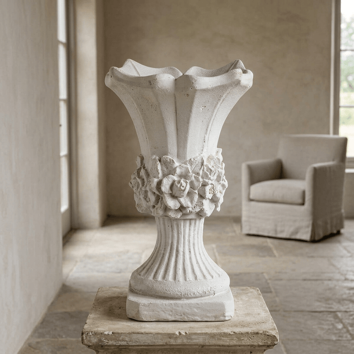 Scalloped floral urn planter displayed empty on a stone plinth in a softly lit neutral sunroom.