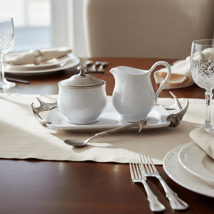 White ceramic sugar bowl and creamer set on a wooden table with silver tray and cutlery.