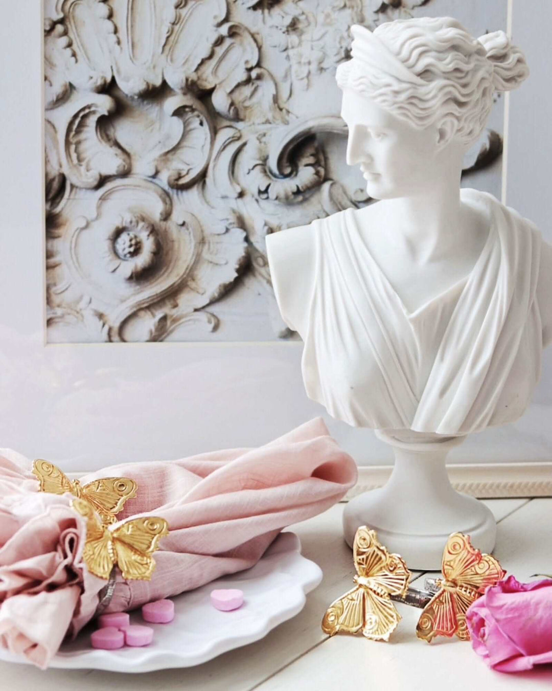 Decorative setting with a classical bust, pink napkin, gold butterfly napkin rings, and pink candies.