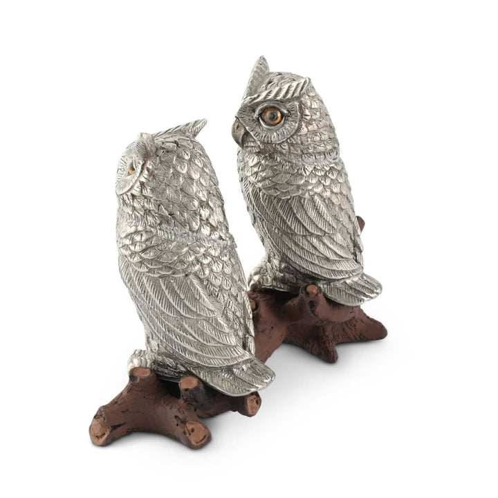 Two silver owl figurines on brown branches against a white background