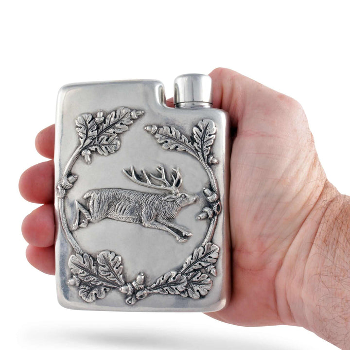 Hand holding a silver flask with deer and leaf design on a white background