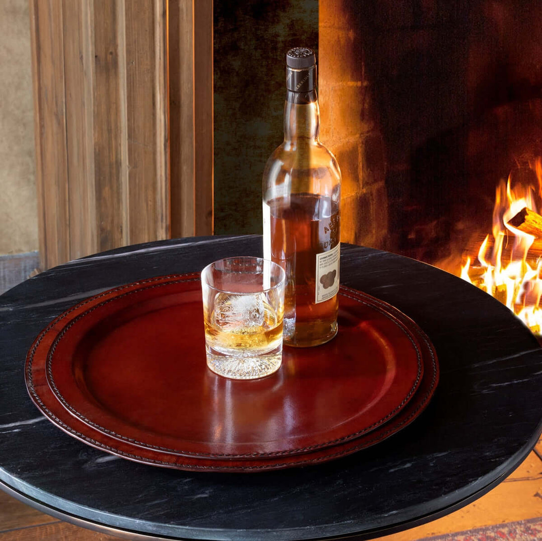 Bottle of whiskey and glass on a leather tray with a fireplace in the background