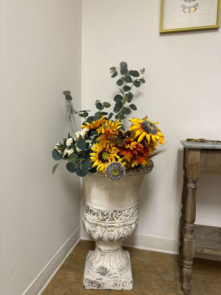 Vintage distressed urn planter filled with vibrant sunflowers and greenery in a cozy indoor setting.