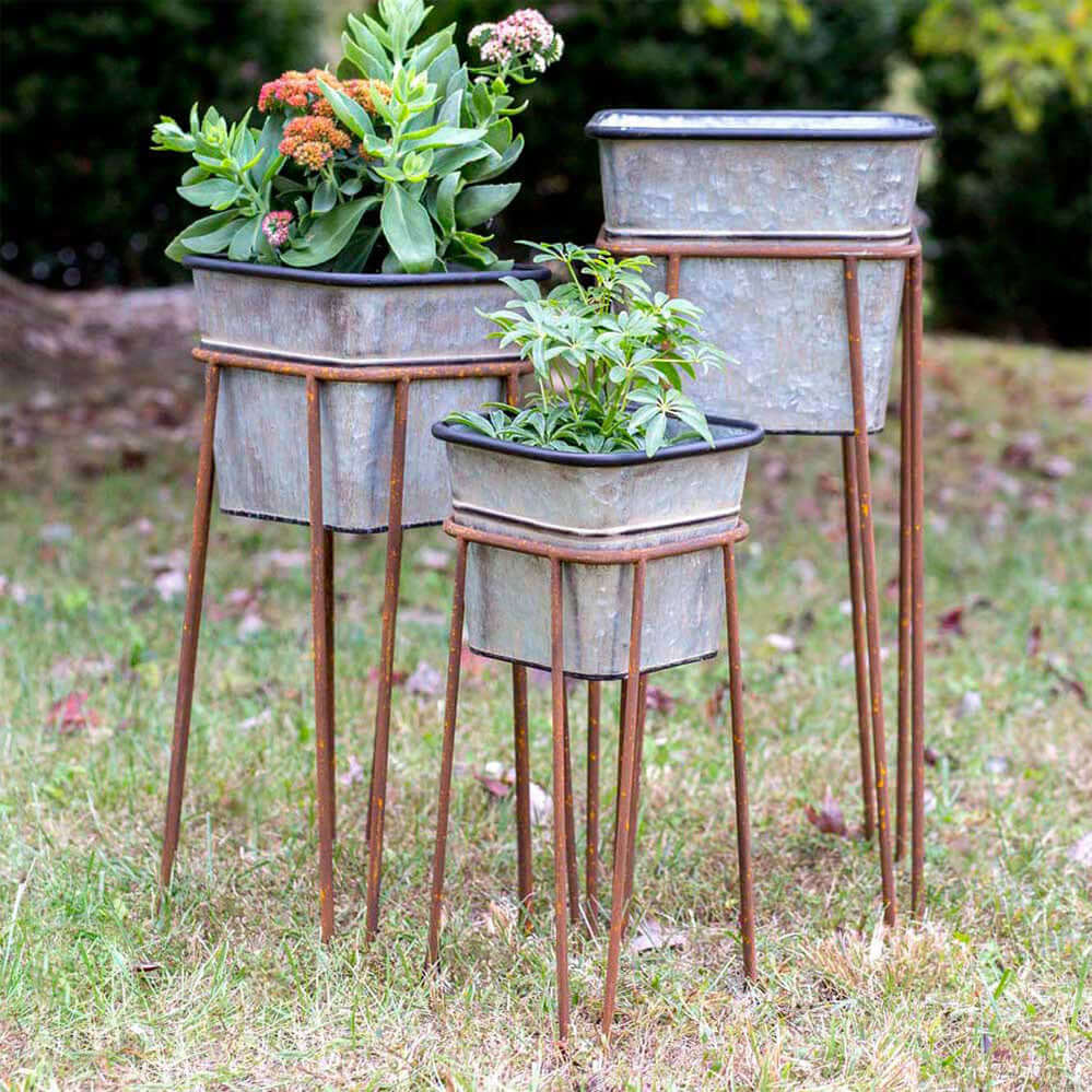 Set of 3 vintage galvanized metal planters with rusted stands, displaying potted greenery in a rustic outdoor setting.