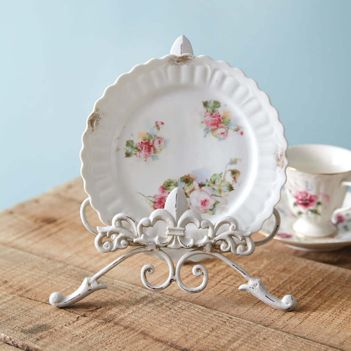 Vintage white metal easel stand displaying a floral decorative plate on a wooden table.