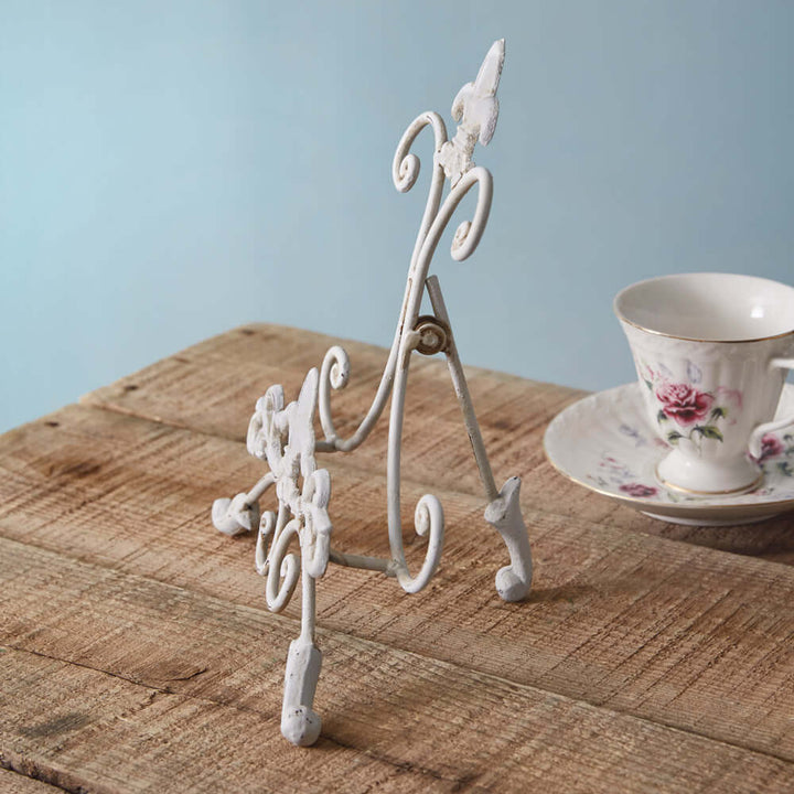 Vintage white metal easel stand with intricate scrollwork on a rustic wooden table, showcasing a decorative teacup.