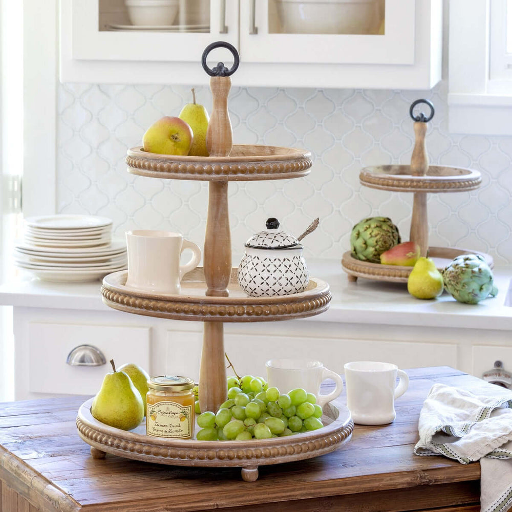 Three-tiered wooden serving tray with fruits and mugs on a kitchen counter.