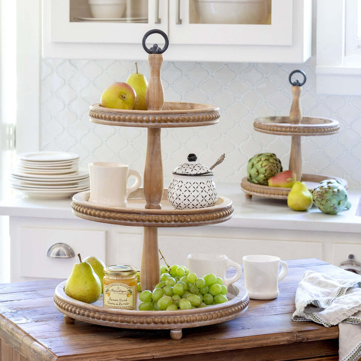 Three-tiered wooden serving tray with fruits and mugs on a kitchen counter.