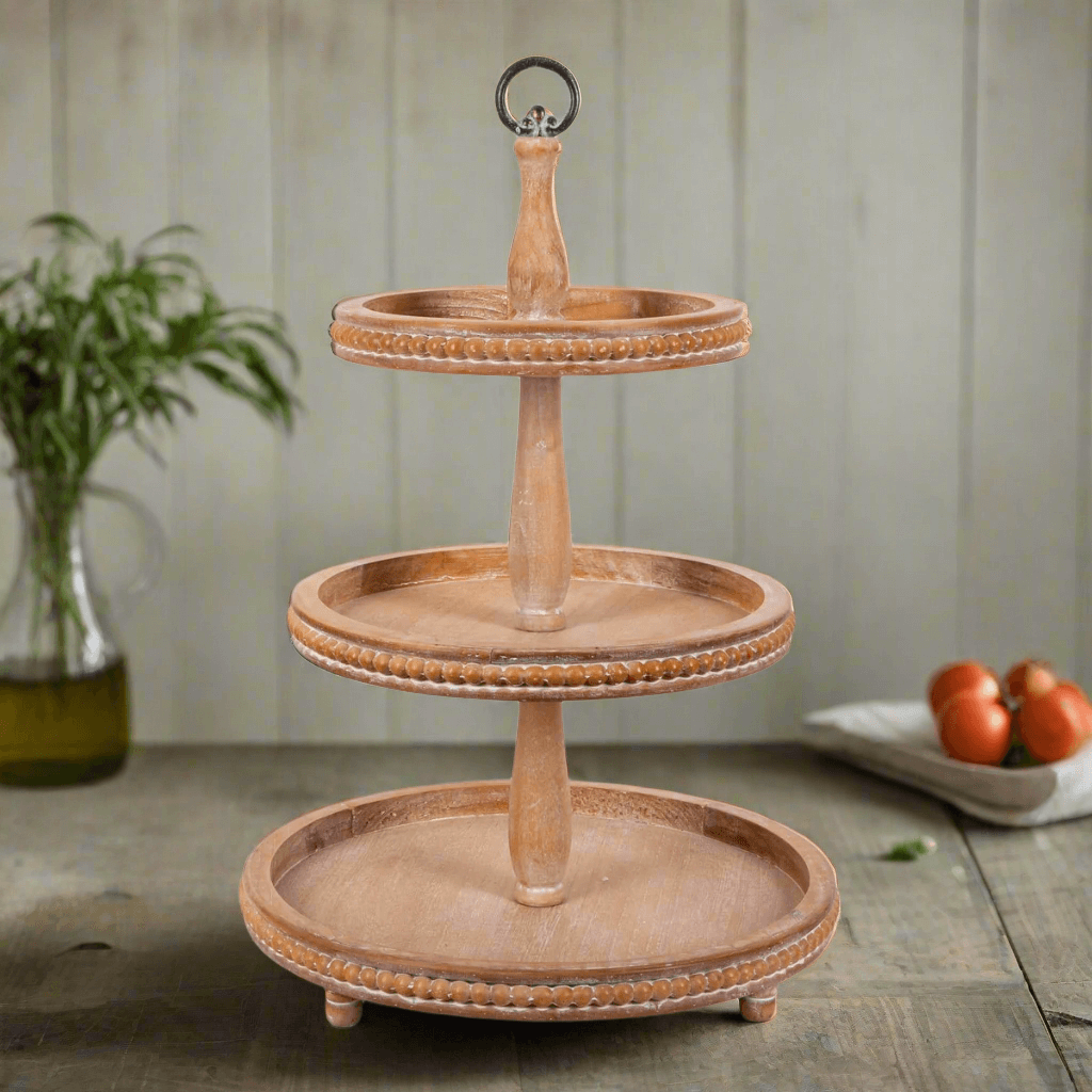 Three-tiered wooden serving tray on a rustic wooden surface with a plant and tomatoes in the background.