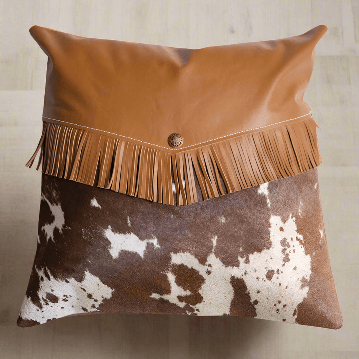 Western cowhide pillow with leather fringe and brass button, adding rustic charm to home decor.