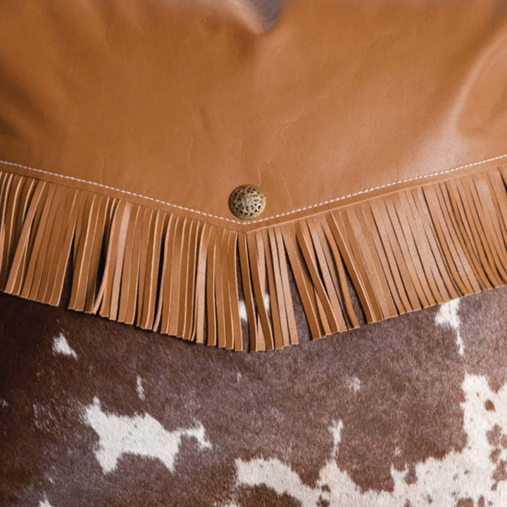 Western cowhide pillow with leather fringe and brass concho button, adding rustic charm to any home decor.