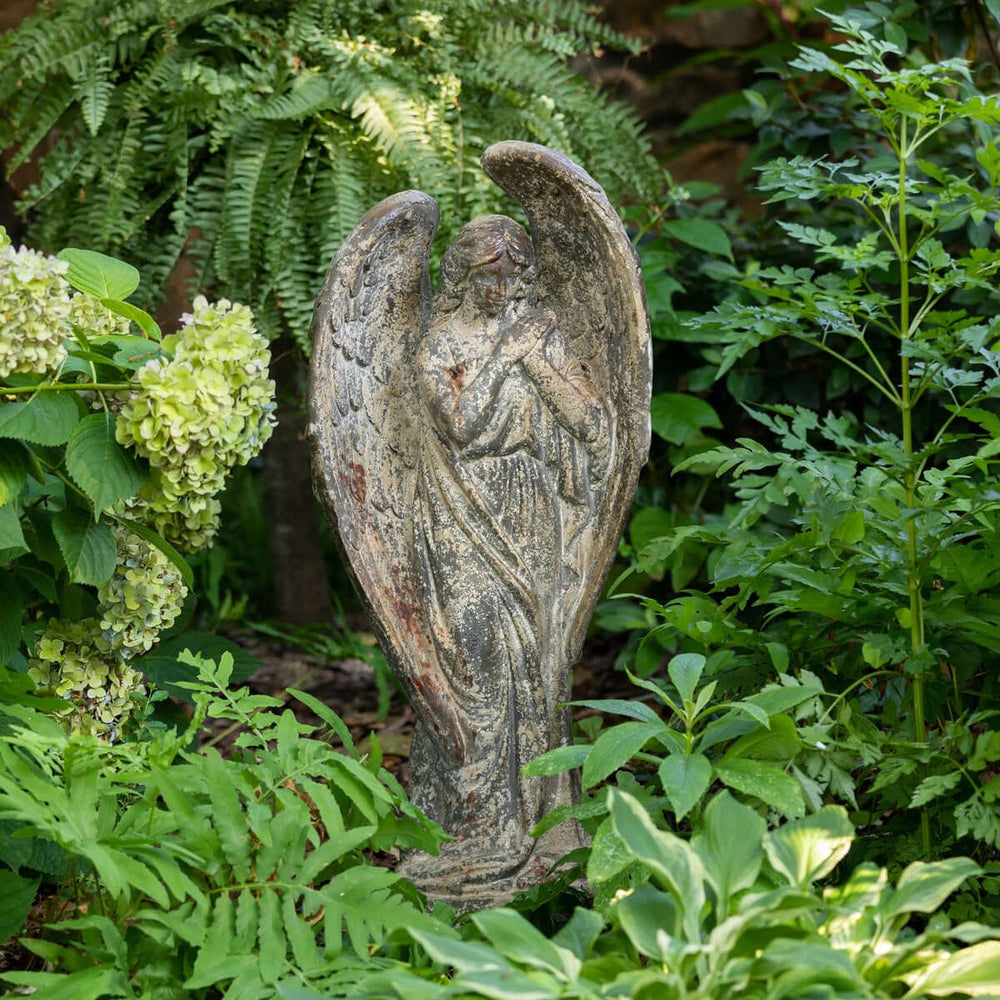 Aged stone Italian garden angel statue with wings, surrounded by lush greenery and flowers in an Old World style garden setting.