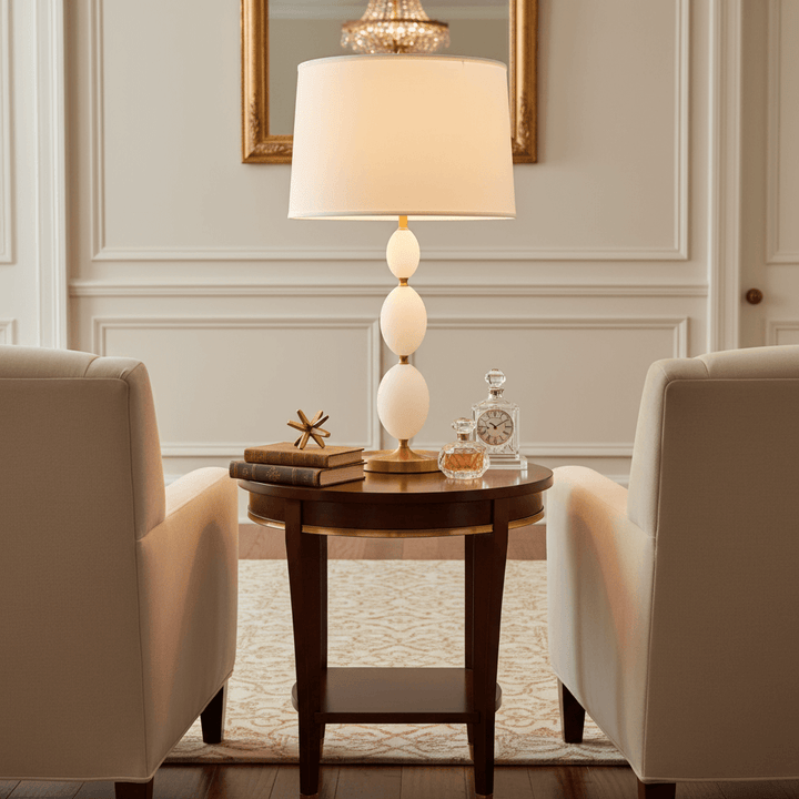Living room with a round wooden table featuring a lamp and decorative items, flanked by two beige armchairs.
