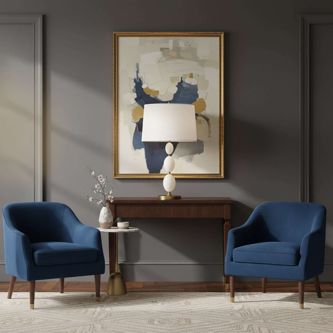 Two blue armchairs in a living room with a abstract painting and lamp on a wooden console.