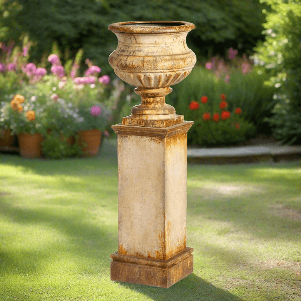 Antique-style iron garden urn with tall pedestal in a garden setting, showcasing a rusted aged patina and fluted design.