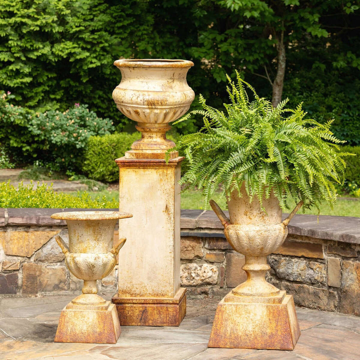Antique-style iron garden urn with tall pedestal in rustic aged patina, displayed outdoors on stone patio with lush greenery.