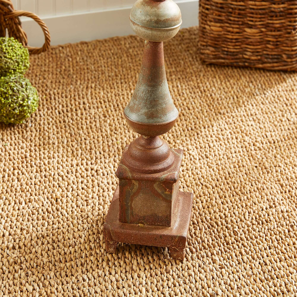 Antique-style rusted metal finial decor on woven mat, featuring a weathered patina and classical architectural design.