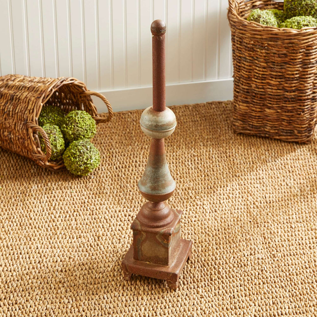 Antique-style rusted metal finial decor on a woven rug, surrounded by rustic baskets and decorative orbs.