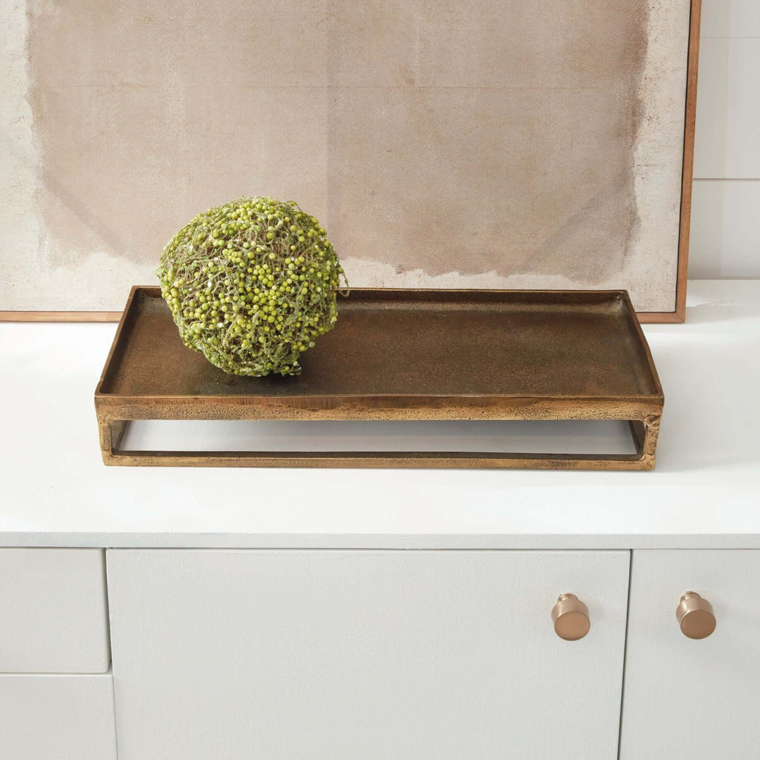 Antique brass raised display tray with a sculptural frame on a countertop, holding a green decorative orb. 