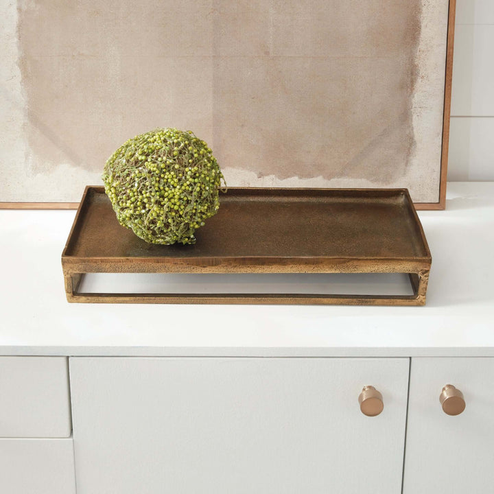 Antique brass raised display tray with a sculptural frame on a countertop, holding a green decorative orb. 