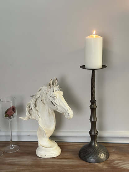 Antique bronze hammered metal candle holder with lit pillar candle, next to ornate white horse sculpture on wooden table.