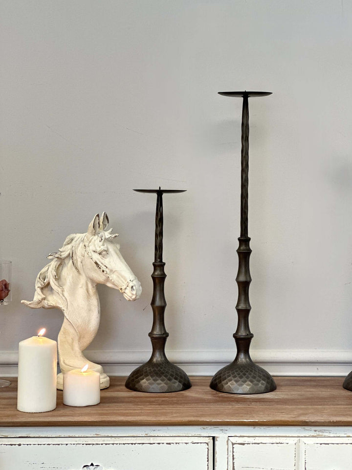 Antique bronze hammered metal candle holders for pillar candles on a wooden surface next to white horse bust decor.