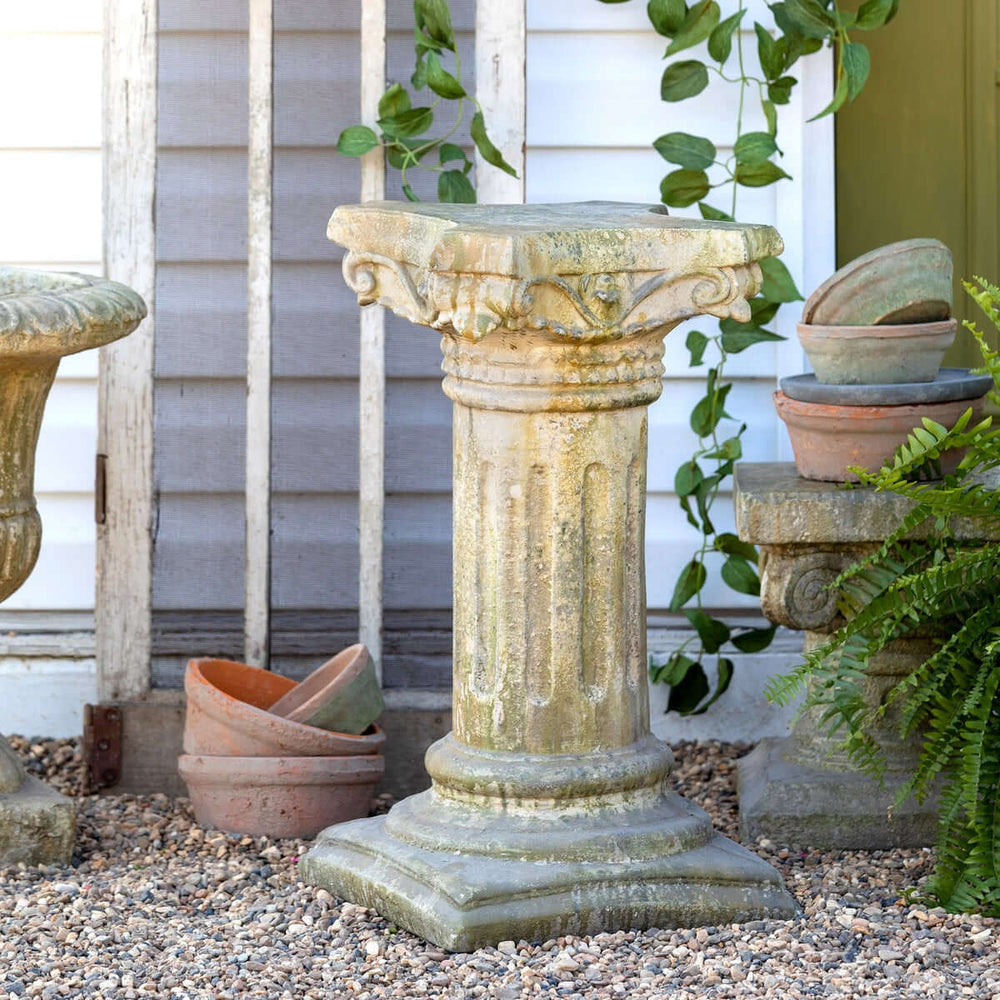 Antique European garden column pedestal with fluted design, topped with cornice, surrounded by pots and greenery in courtyard setting.