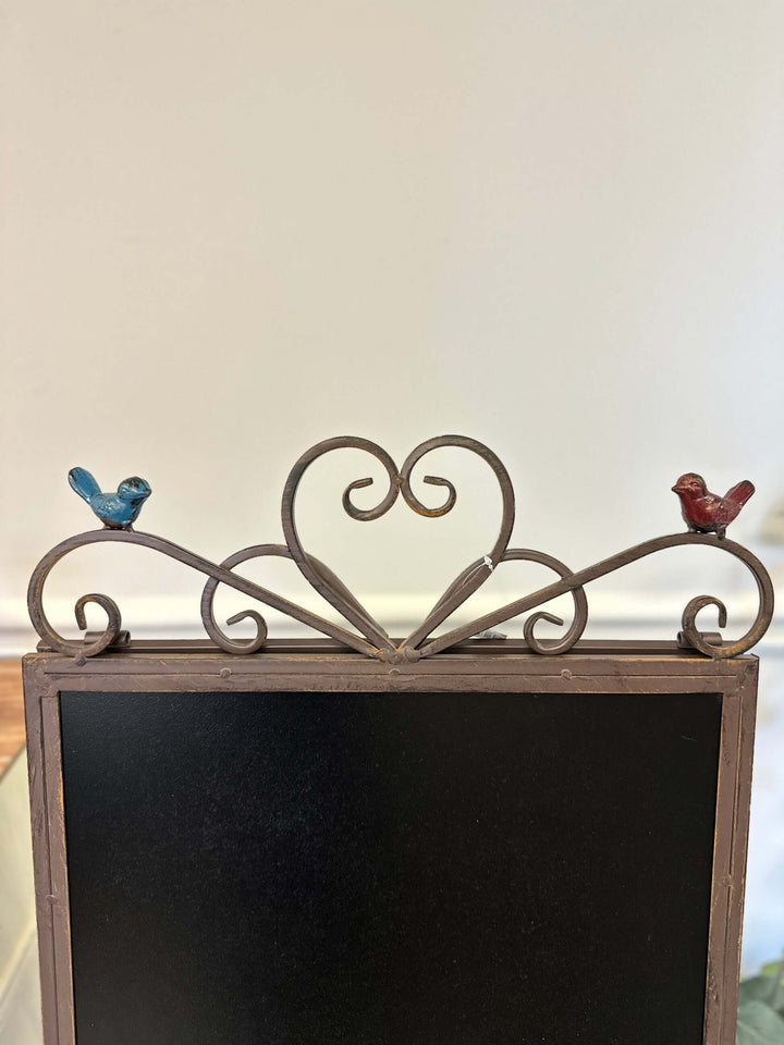 Antique iron chalkboard easel with heart scroll and bird accents, vintage décor piece with matte black writing surface.
