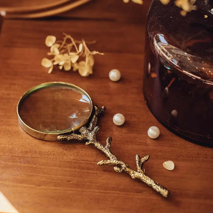 Antique-style gold branch magnifying glass with rustic handle on wooden table, surrounded by pearls and dried flowers.