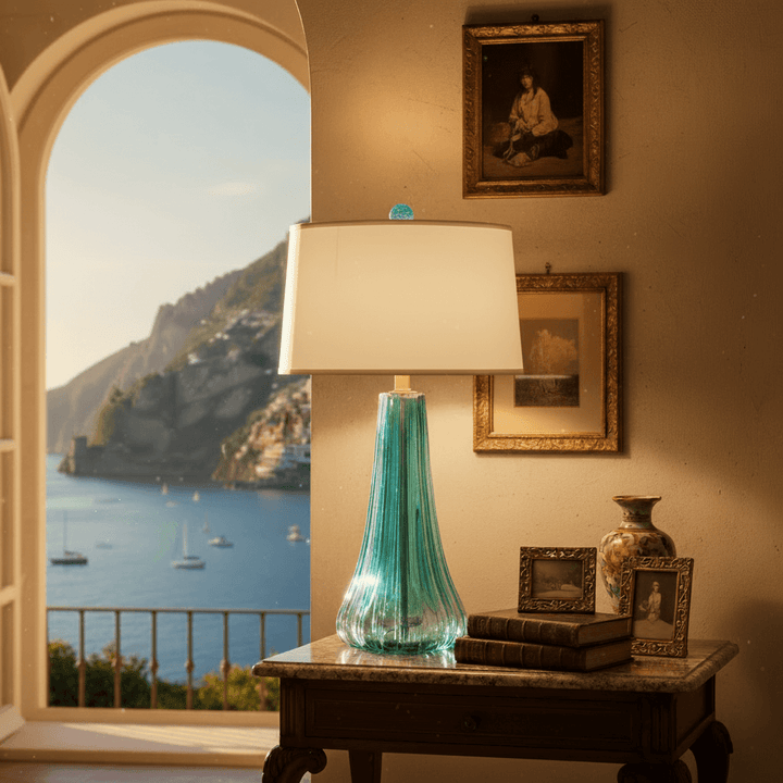 Table lamp with a blue glass base on a table by a window with a scenic Italian coastal view.