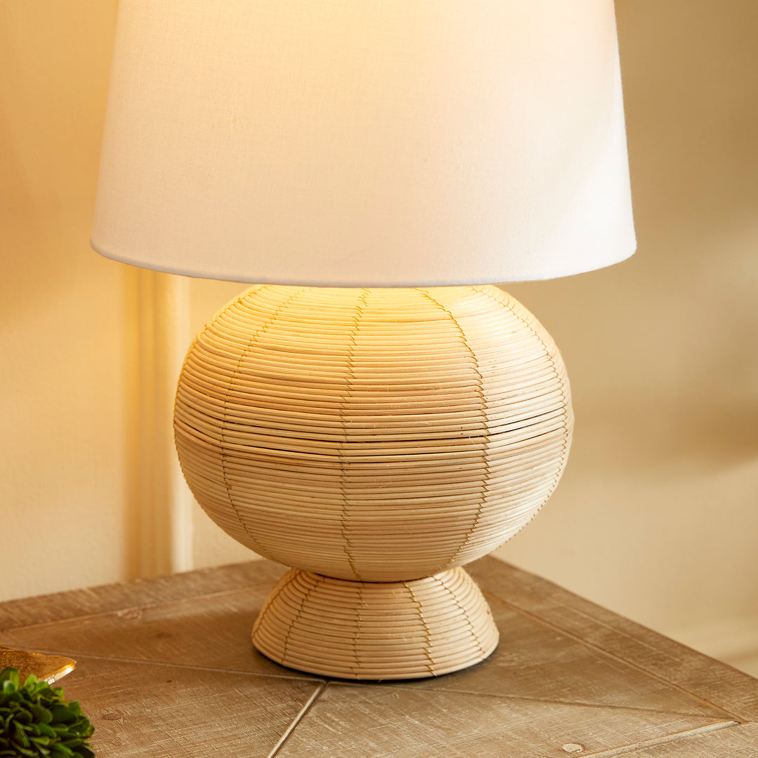 Boho globe rattan lamp with tapered white shade, casting soft, diffused light on a wooden table.