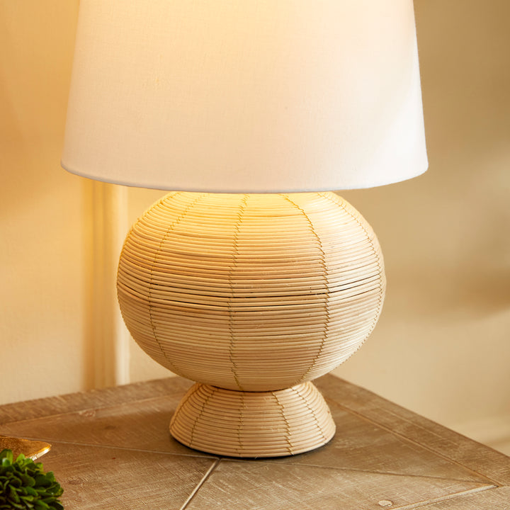 Boho globe rattan lamp with tapered white shade, casting soft, diffused light on a wooden table.