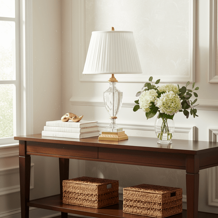 Wooden desk with lamp, books, and flowers in a room with large windows.