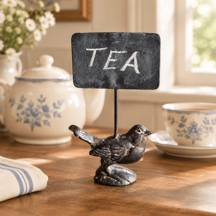 Cast iron bird chalkboard sign labeled Tea on a wooden kitchen counter beside tea accessories.