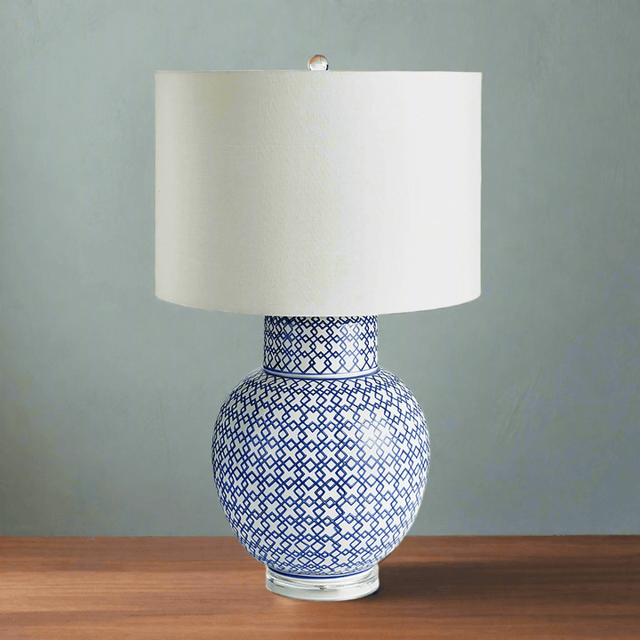 Chinoiserie blue and white ginger jar lamp with a round base and white shade on a wooden table.