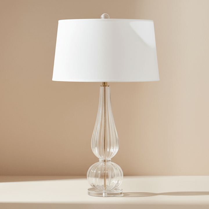 Table lamp with a clear glass base and white shade on a beige surface.