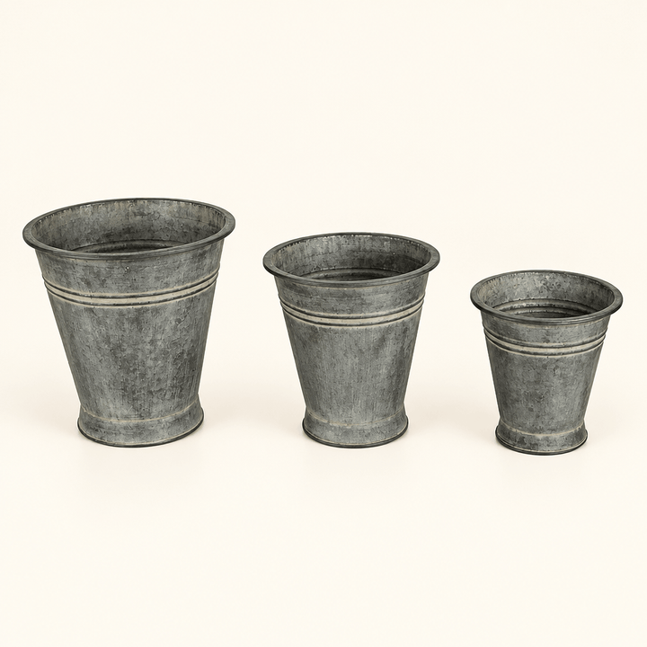 Three graduated metal bucket planters with tapered shapes and an aged finish on ivory.